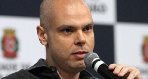 Foto de Bruno Covas, um homem branco, totalmente calvo, de olhos castanhos escuros e sem barba. Na foto, aparece somente a gola de sua camisa social preta. Ele está segurando um microfone próximo à boca. No fundo, vê-se o brasão da prefeitura de São Paulo desfocado.