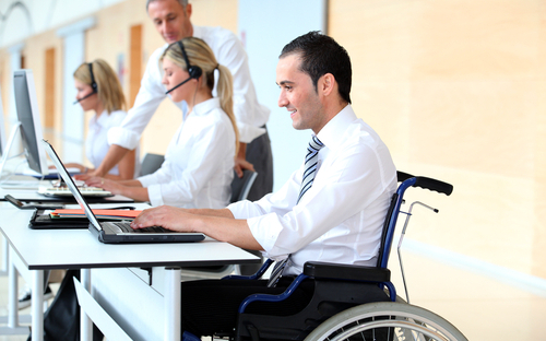 A imagem mostra um homem sentado em uma cadeira de rodas, de frente para um notebook, que está sobre uma mesa branca. Ele é branco, tem cabelos pretos, curtos e lisos, usa uma camisa social branca com uma gravata.