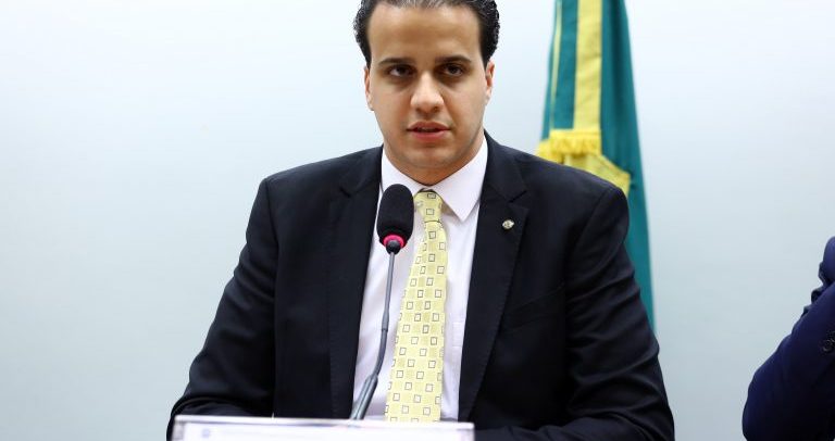 Foto do deputado Lucas Gonzalez. Ele é um homem branco, de cabelos pretos curtos, olhos escuros, veste terno preto, camisa branca e gravata em tom bege. Ele está sentado em uma mesa e em frente a um microfone.
