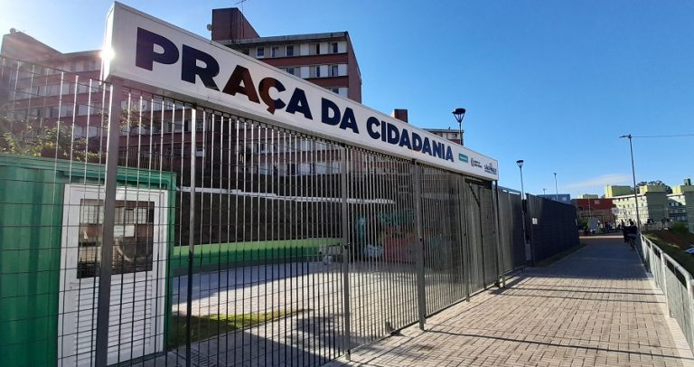 imagem da fachada da Praça da Cidadania, que mostra um portão grande de grades de cor cinza, em um corredor longo com grades em volta.Na parte superior do portão, há uma placa branca onde está escrito “Praça da Cidadania” em letras maiúsculas pretas. Atrás do portão, é possível ver alguns prédios com fachada escura.