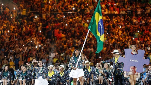 imagem da delegação brasileira no evento de abertura das Olimpíadas Rio-2016. em primeiro plano, a delegação de atletas paralímpicos e uma atleta negra carrega a bandeira do Brasil. Ela veste o uniforme composto por um casaco verde escuro e uma saia curta branca.