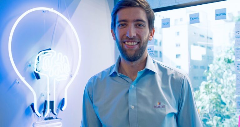 Guilherme Braga, CEO da Egalitê, é um homem branco, de olhos azuis, de cabelos castanhos claros curtos e barba. Ele usa uma camisa social azul clara com o logo da Egalitê do lado esquerdo. Atrás dele, há uma parede no mesmo tom, com o desenho de uma lâmpada acesa em luzes de neon.