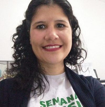 Luciana Sans de Carvalho é uma mulher branca , de cabelos escuros, longos e cacheados, olhos escuros Está vestindo uma camiseta branca com a frase: semana inclusiva, em verde, e um casaco preto. Ela está sorrindo.