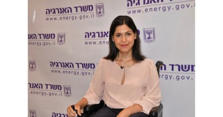 Karine Elharrar é uma mulher branca, de olhos e cabelos escuros e lisos, na altura do ombro. Ela está vestindo uma blusa rosa de mangas compridas e uma calça cinza., e está em uma cadeira de todas. Ao fundo, há uma parede branca com palavras em hebraico.