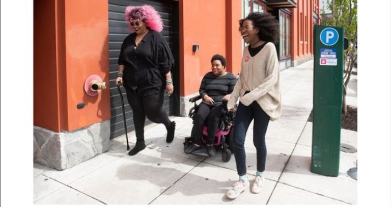 2 mulheres estão na calçada de uma rua. As três são mulheres negras, uma delas usa cadeira de rodas e outra usa uma bengala. A calçada é acessível. Elas parecem conversar e sorriem.