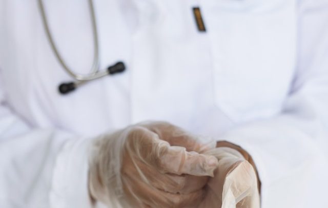 imagem de um médico vestindo jaleco branco e estetoscópio, colocando luvas de plástico. A imagem mostra somente do pescoço para baixo.