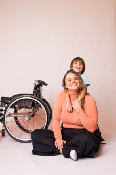 Tábata está sentada no chão, com o filho Francisco com aproximadamente 3 anos de idade. Ela está sentada no chão, os dois estão abraçados e sorriem. A cadeira de rodas está ao lado deles.