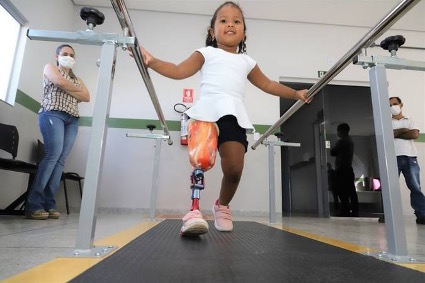 menina de aproximadamente 8 anos de idade caminha com apoio de protese em uma das pernas e segurando em barras de apoio