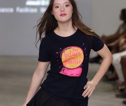 imagem de uma jovem adulta branca, de cabelos longos, lisos e castanhos. ela desfila em uma passarela de moda e faz uma pose para a foto. ela tem síndrome de down