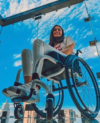 Uma jovem mulher sorrindo, sentada em uma cadeira de rodas que está sendo içada e suspensa no ar. Ela está em um ambiente de paredes e teto envidraçados, pelos quais se vê o céu azul com nuvens brancas