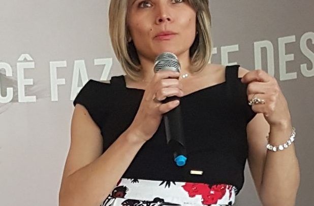 Marinalva Cruz é uma mulher branca, de olhos castanhos, cabelos lisos na altura dos ombros, usa blusa preta e saia branca com estampa florida. Está falando ao microfone. É uma mulher com deficiência física.