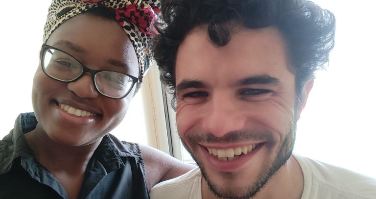 Lucas é um homem jovem, de pele branca, cabelos encaracolados marrom escuro, usa barba pequena e está sorrindo. Vanessa é uma jovem de pele negras, usa turbante colorido cobrindo seus cabelos, usa óculos com armação marrom, e está sorrindo. Os dois estão lado a lado, abraçados.