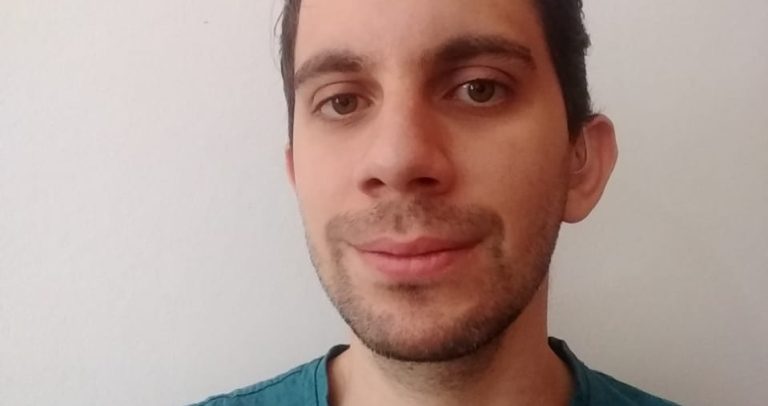Andrey Marcondes é um homem branco, com cabelos curtos marrons e olhos marrons, barba e bigodes bem curtos, e está sorrindo. Ele usa uma camiseta azul petróleo.