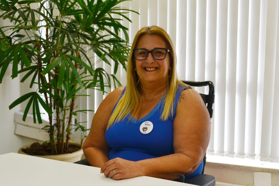 Marly é uma mulher branca, de cabelos lisos e loiros um pouco abaixo dos ombros. Usa uma blusa azul sem mangas, óculos de grau e cadeira de rodas. Está sorrindo.