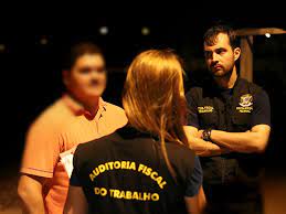 foto de três pessoas, uma mulher de costas e dois homens de frente, com a imagem do rosto borrada. A mulher e um dos homens usam coletes pretos, com texto nas costas, onde se lê: Auditoria Fiscal do Trabalho.