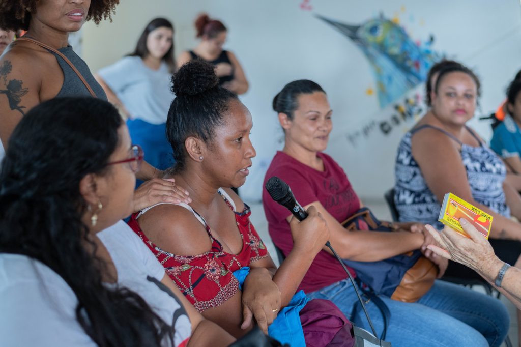 Grupo de mulheres sentadas em cadeiras tipo auditório, com destaque para 4 delas que estão na fileira da frente. Uma delas é uma mulher negra está falando ao microfone., que segura na mão direita.
