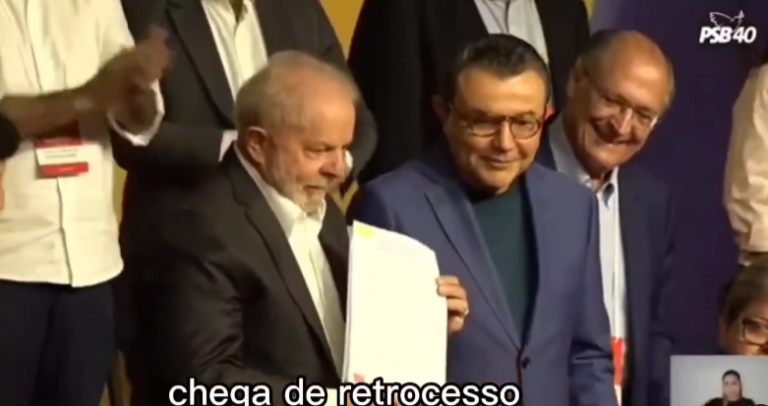 homem branco, de cabelos brancos, veste um terno preto com uma camisa branca por baixo. ele segura em suas mãos uma folha. ao seu lado estão dois homens brancos sorrindo. na foto está a legenda: chega de retrocesso. no canto inferior direito está uma intérprete de Libras