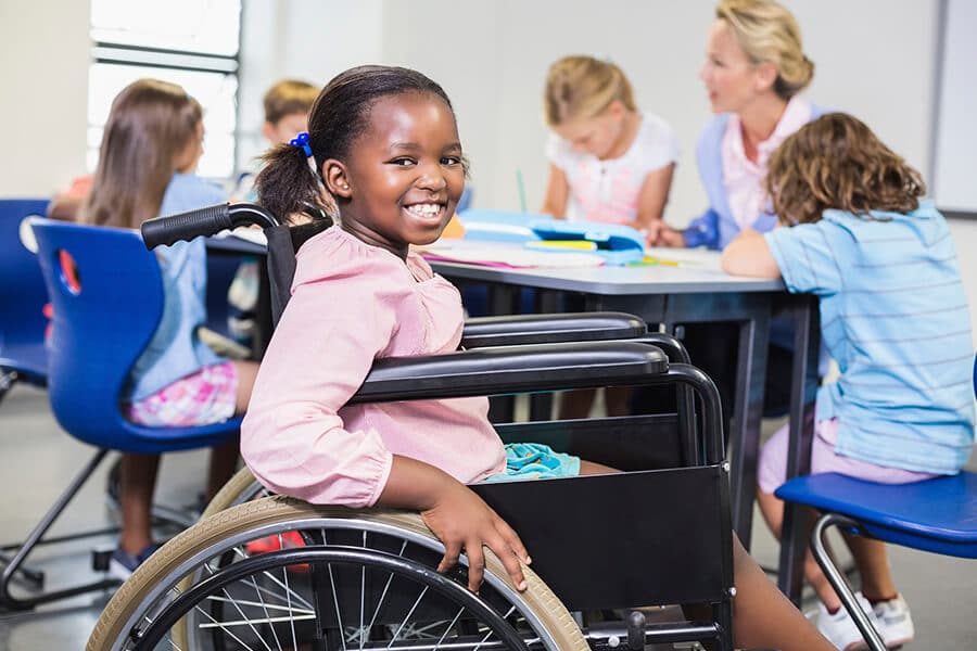 Uma criança negra sorrindo, ela usa uma cadeira de rodas e está com uma blusa rosa e short azul. No fundo tem ambiente escolar, com algumas crianças ao redor de uma mesa junto com a professora.