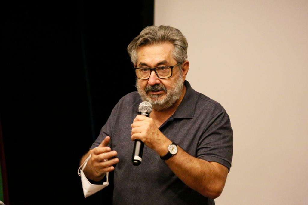 Foto do Dr. Kal. Ele tem os cabelos, barba e bigode grisalhos, usa blusa cinza escuro, está de pé falando ao microfone.
