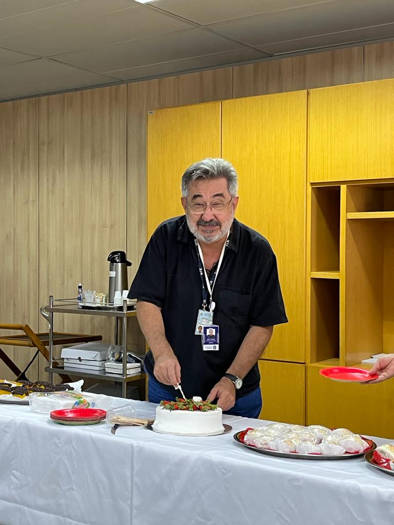 Kal está de pé, usa camisa preta e calça jeans, e está sorrindo e cortando o bolo de aniversário, que é branco com morangos no topo.