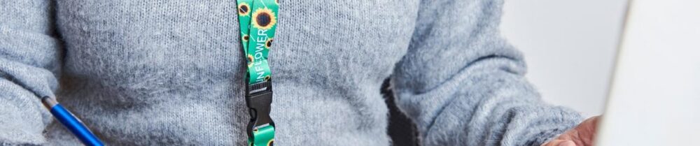 foto do tronco de uma pessoa, que usa blusa de lã em cor cinza claro e cordão azul claro com girassóis amarelos. A pessoa está diante de um notebook e segura um lápis na mão.