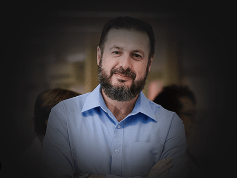 Flávio Gonzales é um homem branco, com olhos, cabelo, barba e bigode de cor marrom escuro. Usa uma camise azul clara, e está com leve sorriso.
