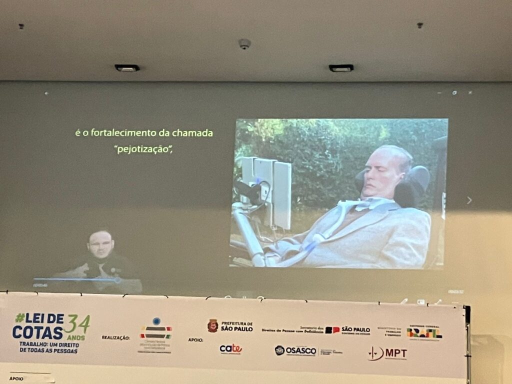 Rick Ribeiro está deitado em uma cadeira de rodas mecanizada, com a cabeça apoiada e um tudo de ventilação em torno do pescoço. Ele lê um tablet fixado na frente da sua cabeça. Ele é branco, grisalho, usa óculos e está vestindo paletó cinza claro e camisa azul claro e colete de lã cinza escuro.
