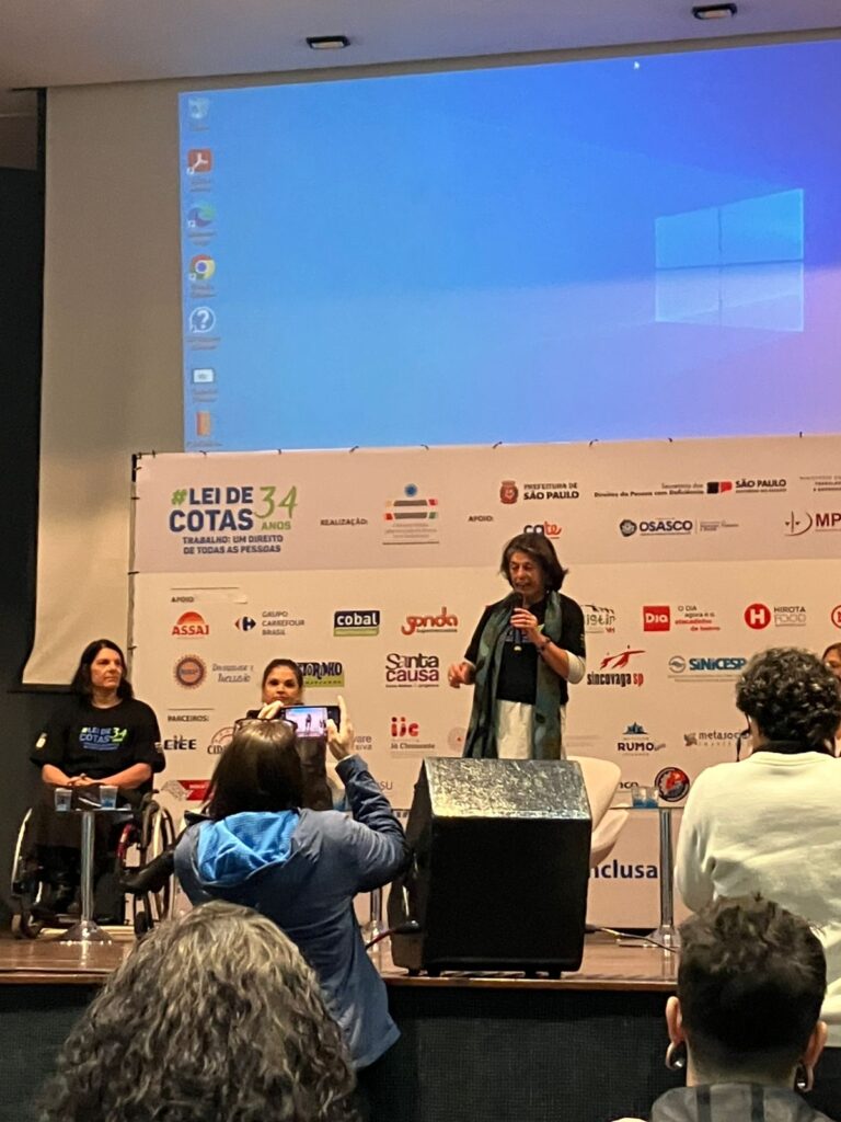 Silvana Gimenes está falando ao microfone de pé no palco, ao lado de Dra Ruth e da secretária Anna Paula Feminella.