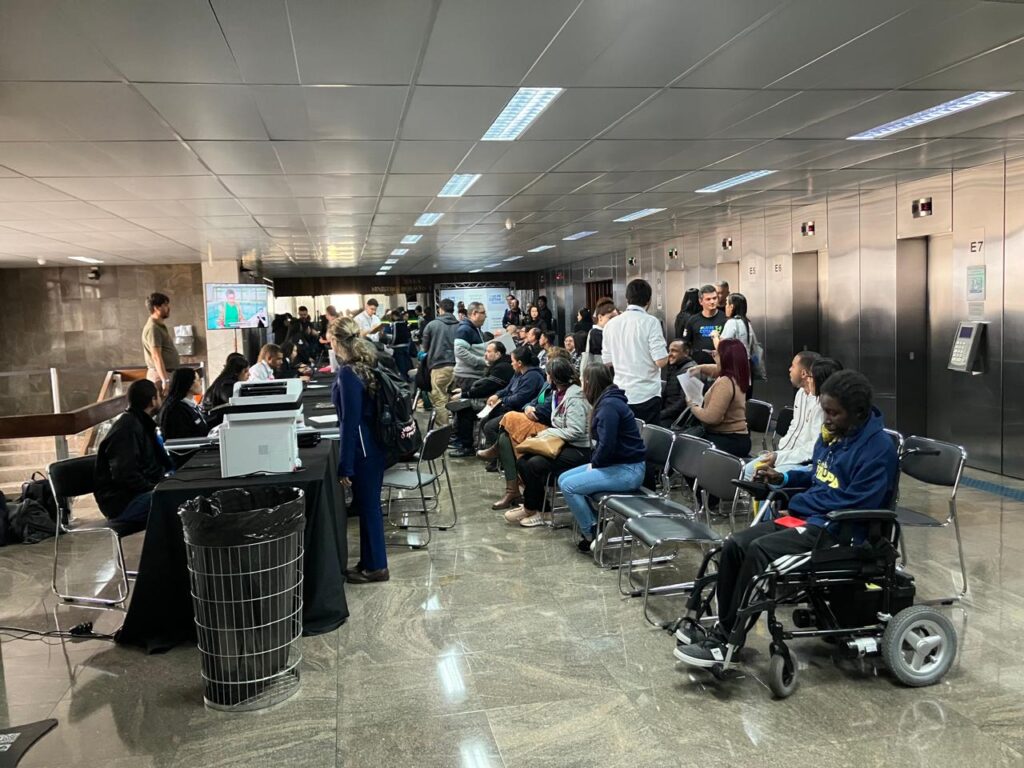 foto mostra o ambiente de cadastro e de entrevistas dos candidatos. Há mesas com atendentes em frente a cadeiras onde as pessoas esperam pelo atendimento. O ambiente está cheio de pessoas sendo atendidas e aguardando o atendimento.