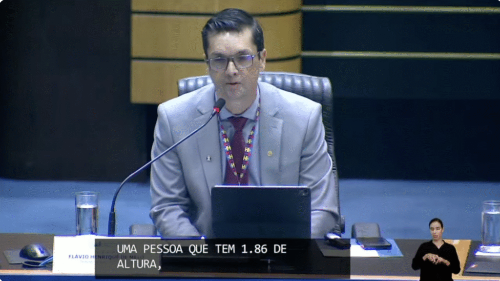 Dr. Flavio Henrique de Melo é um homem de pele branca, cabelo castanho escuro, usa óculos, paletó e camisa azul claro, gravata vinho e o crachá de identificação de pessoas autistas.