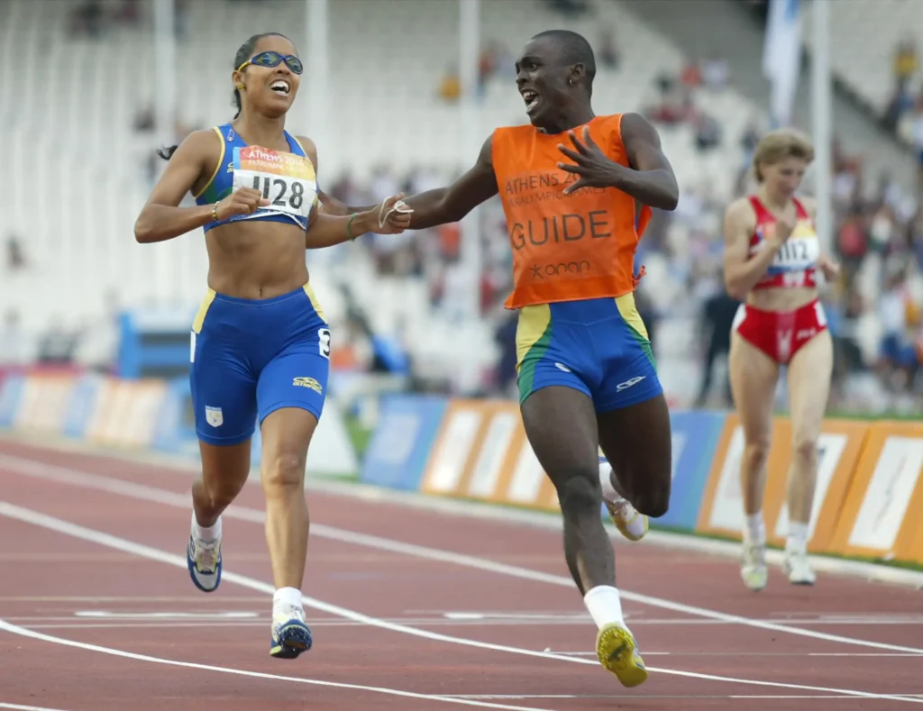 Adria Santos é uma mulher de pele negra, cabelos pretos presos na nuca. Usa top e shorts de corrida, nas cores azul, verde e amarelo, com um número de corredora no peito. Está correndo em uma pista, usa óculos escuros e está sorrindo. Ao seu lado, tocando seu antebraço, seu treinador está correndo e sorrindo. É um homem negro, de cabelos raspados, com músculos bem definidos. Usa shorts azul e camiseta laranja.