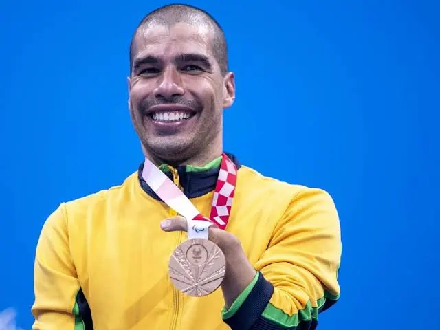 Daniel Dias é um homem negro, de olhos castanhos e cabelos raspados. Na foto usa camiseta amarela com detalhes em verde e preto, está sorridente  mostrando a medalha de ouro que está pendurada no pescoço.