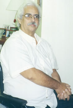 Foto de Cândido Pinto de Melo, onde aparece da cintura para cima. Homem de pele e cabelos brancos, usa óculo de grau, bigode grisalho, e está vestindo camisa branca. Está sentado.