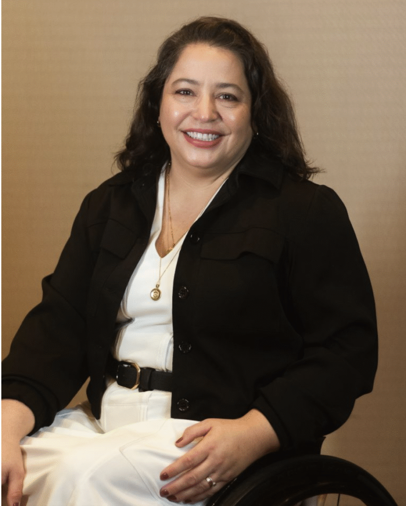 Carolina é uma mulher branca, de cabelo liso, marrom, cortado abaixo dos ombros. Usa vestido branco e casaco preto. Está sentada na cadeira de rodas e sorri.