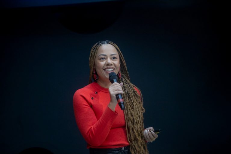 Ana é uma mulher negra, de cabelo com tranças longas, usa casaco vermelho, está falando ao microfone e sorrindo.