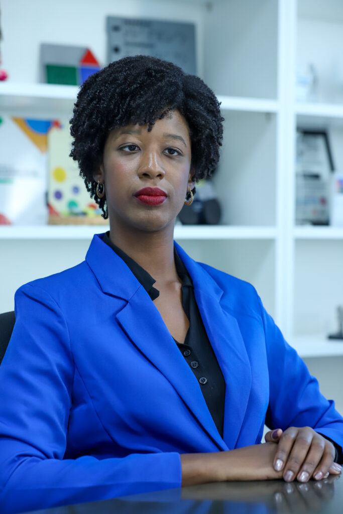 A secretária nacional é uma mulher de pele e olhos negros, cabelo preto crespo curto com cachinhos definidos, usa paletó azul cobalto sobre uma blusa preta, brincos pequenos, batom vermelho. Está sentada, em ambiente de escritório, com as mãos sobre a mesa, em uma pose elegante.