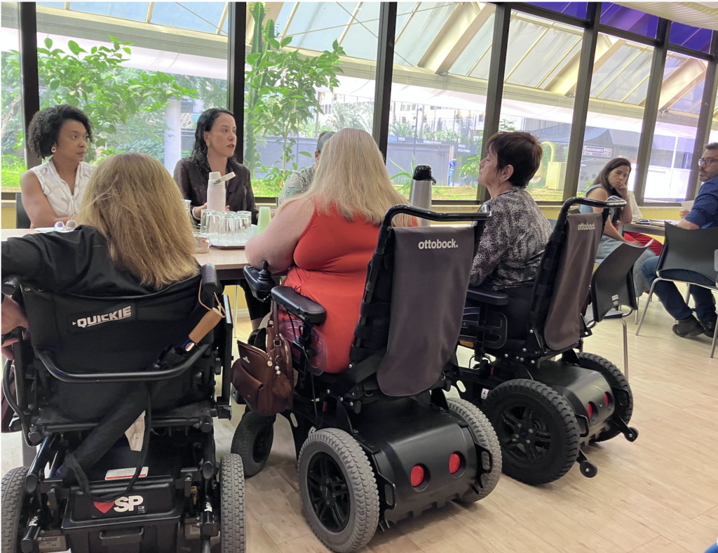 Grupo de integrantes do Fórum reunido. Todos estão trabalhando em grupo. São três mesas compridas dispostas em U na sala. Algumas pessoas estão em cadeira de rodas.