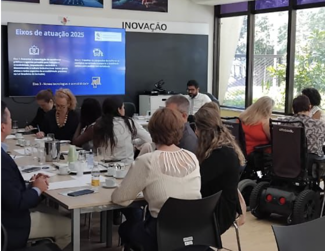 Grupo de integrantes do Fórum reunido. Todos estão trabalhando em grupo. São três mesas compridas dispostas em U na sala.