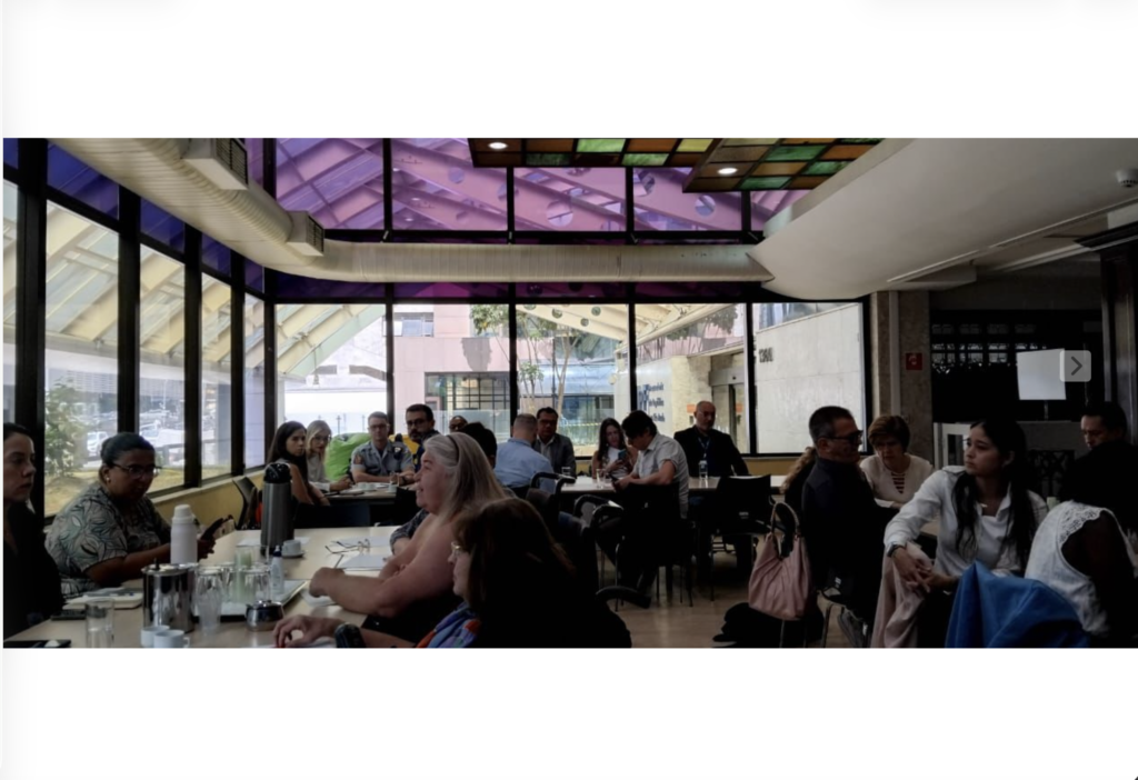 Grupo de integrantes do Fórum reunido. Todos estão trabalhando em grupo. São três mesas compridas dispostas em U na sala.