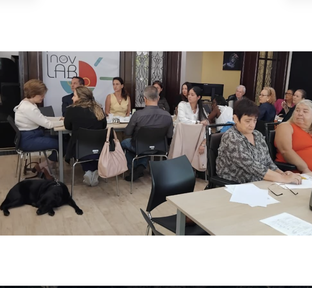 Grupo de integrantes do Fórum reunido. Todos estão trabalhando em grupo. São três mesas compridas dispostas em U na sala.