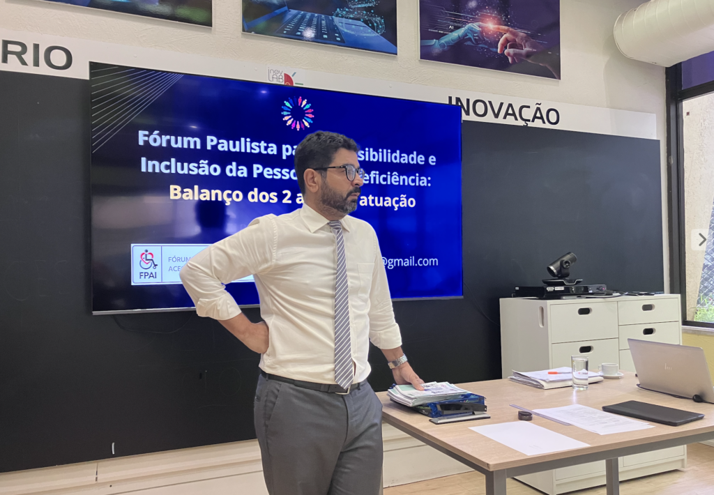 Dr José Rubens Plates é um homem de pele branca, cabelo, olhos e barba marrons, usa óculos de grau. Na foto está de pé à frente, conduzindo a reunião do Fórum. Ele usa calças e gravata cinzas e uma camisa de mangas compridas de cor branca.