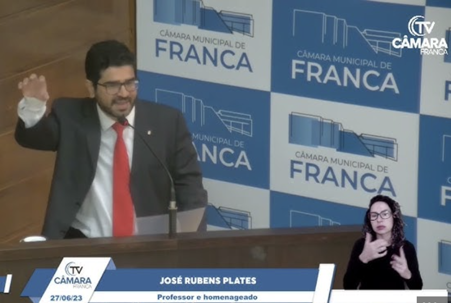 Print da transmissão da homenagem recebida por Dr José Rubens Plates pela Câmara Municipal de Franca, em 2023. Ele  está de pé falando ao microfone, usa paletó cinza escuro, camisa branca e gravata vermelha. Ao fundo aparece um banner escrito Câmara Municipal de Franca com ilustrações em azul e branco. No canto inferior direito aparece a intérprete de Libras, usando roupas pretas.