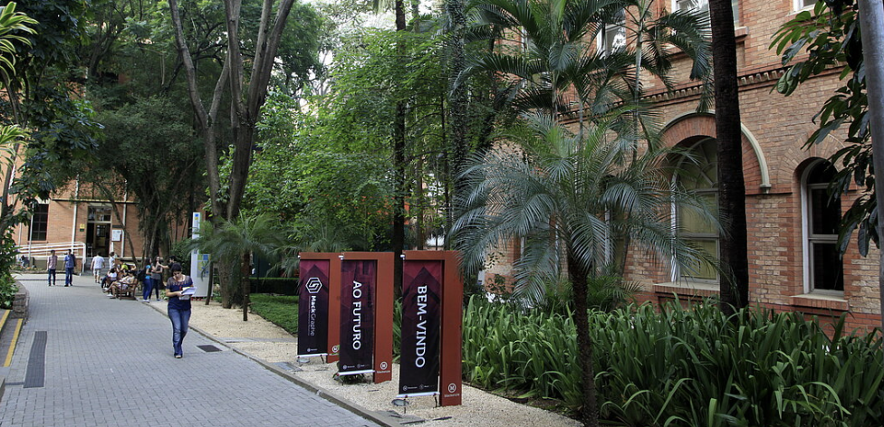 Vista de uma alameda interna da Universidade Mackenzie em Higienópolis. O ambiente é bastante arborizado, com construções antigas bem preservadas, com tijolos à vista e calçamento de pedras.