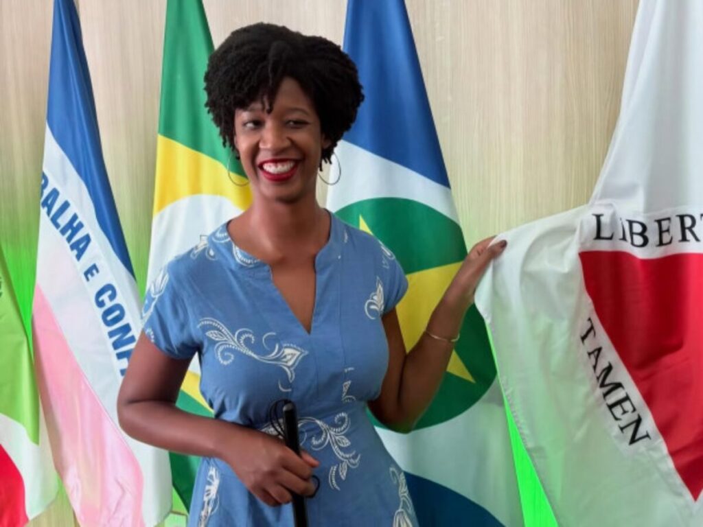 A secretária nacional é uma mulher de pele e olhos negros, cabelo preto crespo curto com cachinhos definidos, usa blusa azul claro, brincos pequenos de argola, batom vermelho. Está de pé, em ambiente de escritório, sorrindo e segurando a ponta da Bandeira de Minas Gerais. Ao fundo aparecem outras bandeiras oficiais.