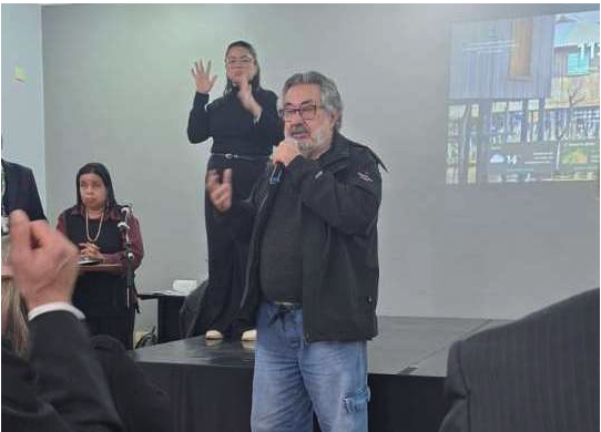 Dr. José Carlos do Carmo fala ao microfone, de pé. Ele usa casado preto e calça jeans. No palco aparece também uma intérprete de Libras.